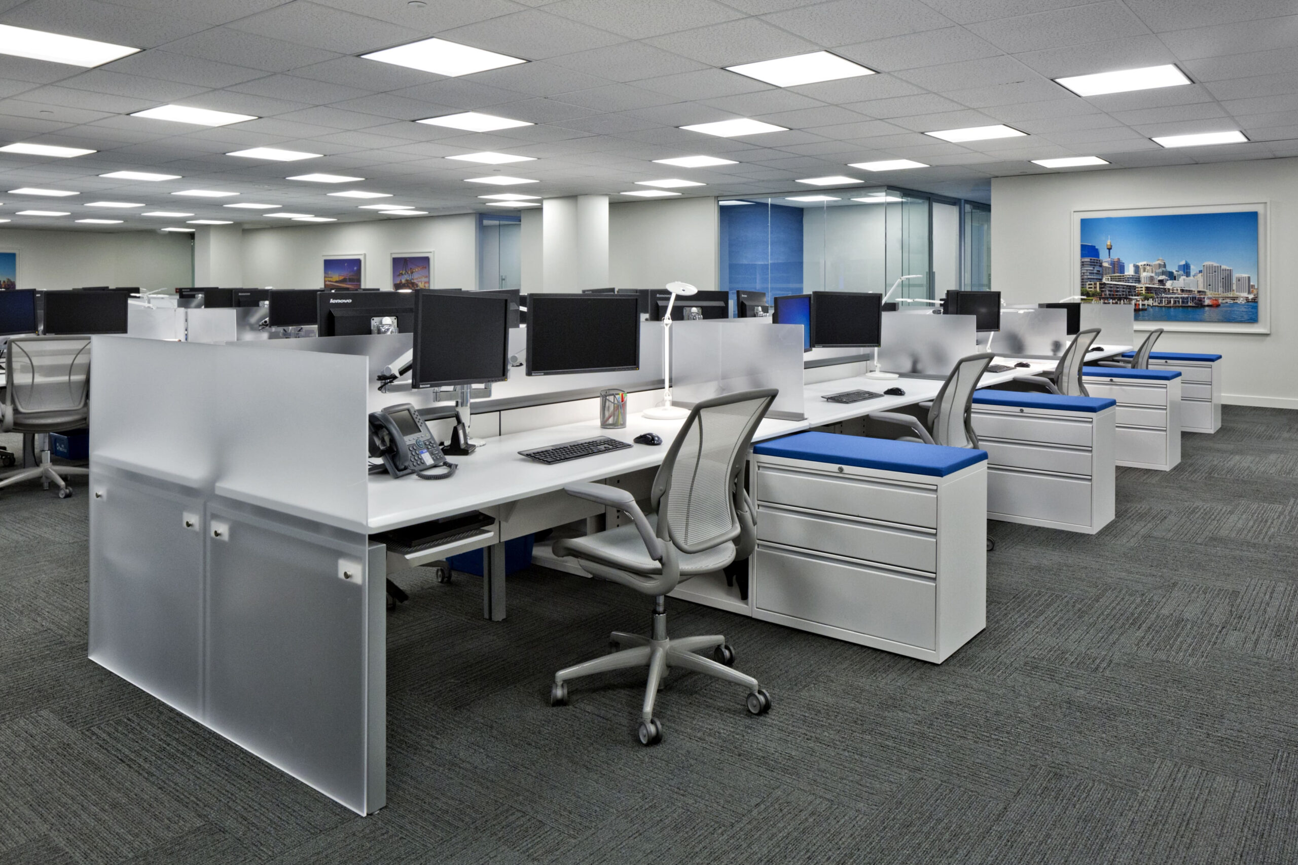 Spacious, modern open-plan office with rows of white desks, ergonomic chairs, and computers. Bright lighting, large windows, and wall art create a clean, professional atmosphere.