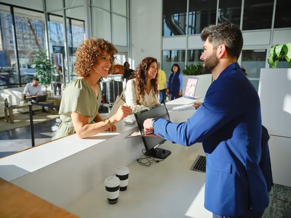A man in a blue suit assists two smiling women at a bright, modern reception desk. Sunlight streams in, creating a welcoming atmosphere.