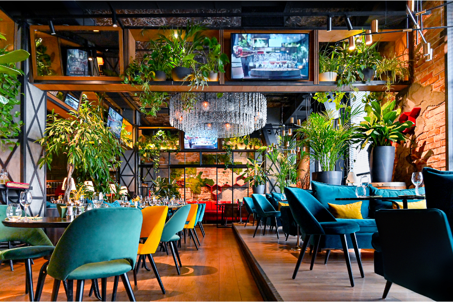 Chic restaurant with vibrant teal and yellow chairs, lush plants, and a crystal chandelier. Exposed brick walls and hanging TVs create a modern, cozy ambiance.